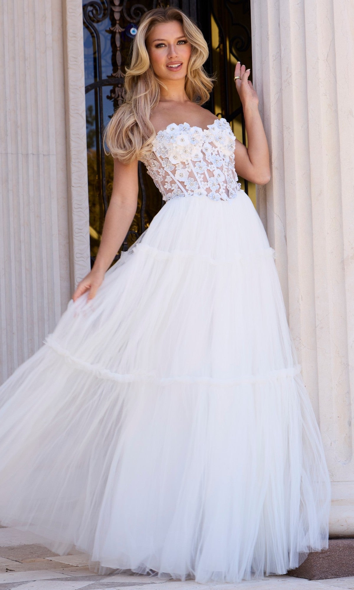 A woman wearing the Jovani 45866 Long Prom Dress in off white, featuring a strapless floral-embellished sheer corset bodice and flowing tulle A-line skirt, stands in front of a columned entrance.